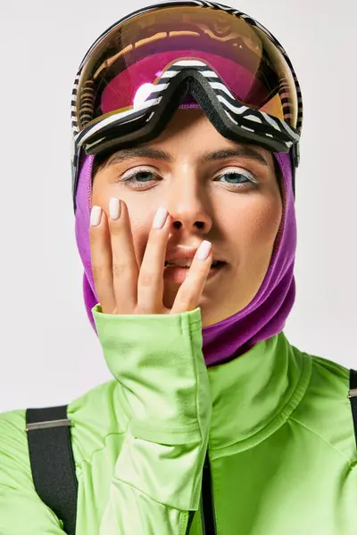 Mujer joven en ropa de esquí de invierno con pasamontañas en la cabeza posando con las manos cerca de la cara en gris - foto de stock
