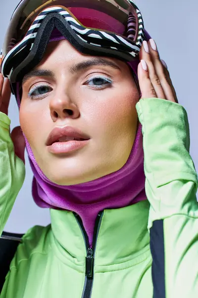 Jeune femme en hiver port actif avec cagoule sur la tête portant des lunettes de ski sur fond gris — Photo de stock