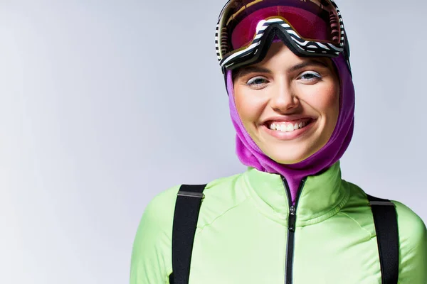 Mujer alegre en invierno ropa activa con pasamontañas en la cabeza y gafas de esquí sobre fondo gris - foto de stock