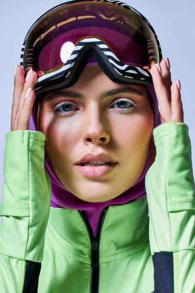 Bonita mujer en invierno ropa activa con pasamontañas en la cabeza ajustando gafas de esquí sobre fondo gris - foto de stock