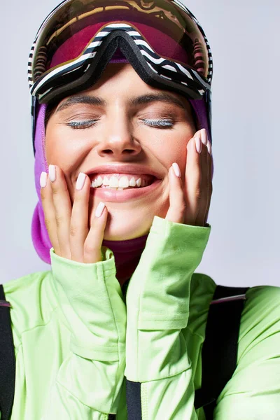 Portrait de femme joyeuse en vêtements de ski d'hiver avec cagoule sur la tête touchant visage sur gris — Photo de stock