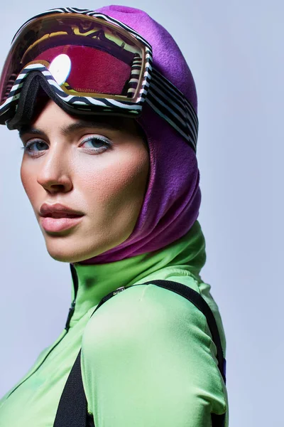 Retrato de mujer en ropa de esquí con pasamontañas en la cabeza mirando a la cámara en gris, maquillaje de invierno - foto de stock