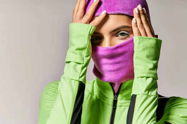 Retrato de mujer deportiva con ojos azules usando máscara de esquí púrpura y mirando a la cámara en gris - foto de stock