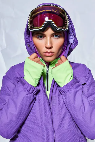 Mujer seria con ojos azules posando en googles de esquí, pasamontañas y chaqueta de invierno sobre fondo gris - foto de stock