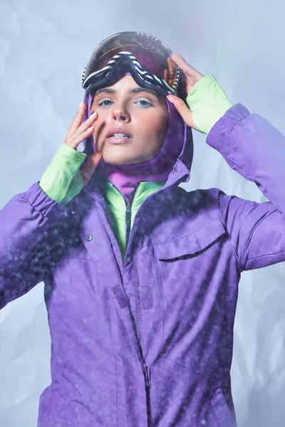Belle femme en cagoule et veste d'hiver portant des lunettes de ski sur fond gris, journée enneigée — Photo de stock