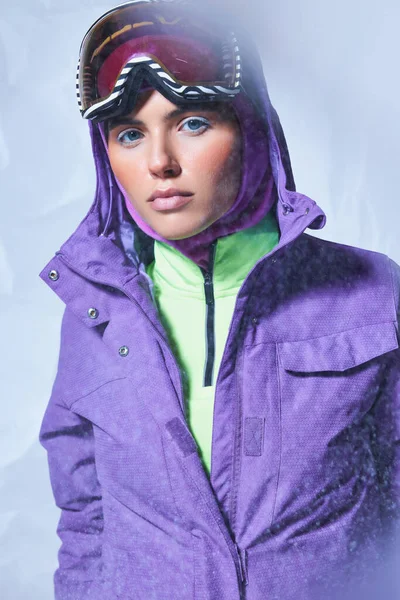 Mulher atraente em balaclava, casaco de inverno roxo e googles de esqui posando em cinza, nevando dia — Fotografia de Stock