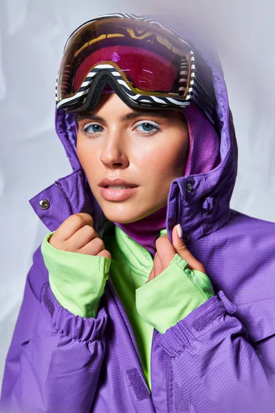 Atractiva mujer en pasamontañas y esquí googles posando en chaqueta de invierno elegante y púrpura en gris - foto de stock