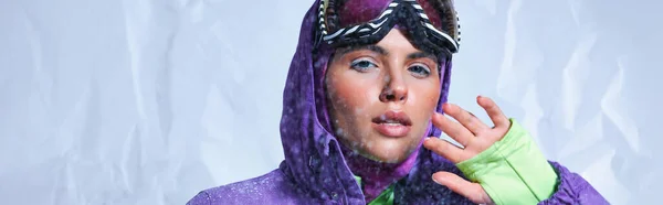Jolie femme en cagoule, veste d'hiver violette et googles de ski posant sur gris, bannière enneigée — Photo de stock