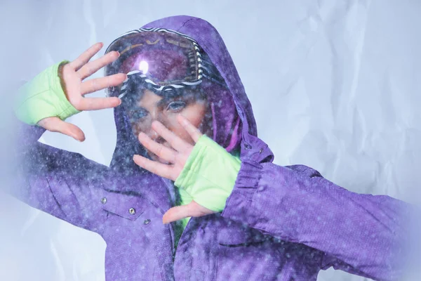 Atractiva mujer en pasamontañas, chaqueta de invierno púrpura y esquí googles posando en gris, clima nevado - foto de stock