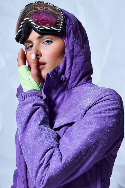 Atractiva mujer en pasamontañas y esquí googles sobre fondo gris, nieve sobre chaqueta de invierno púrpura - foto de stock