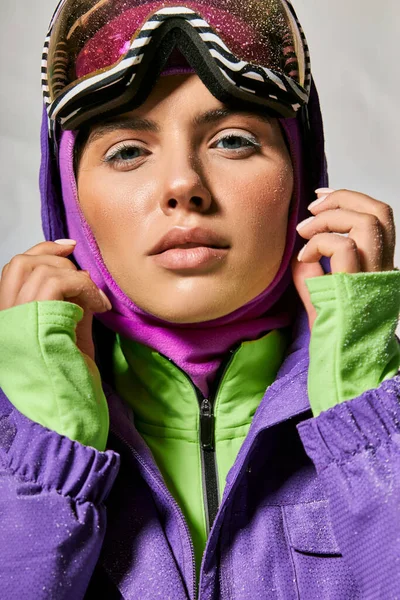 Clima frío, hermosa mujer en pasamontañas y esquí googles posando en chaqueta de invierno púrpura sobre gris - foto de stock