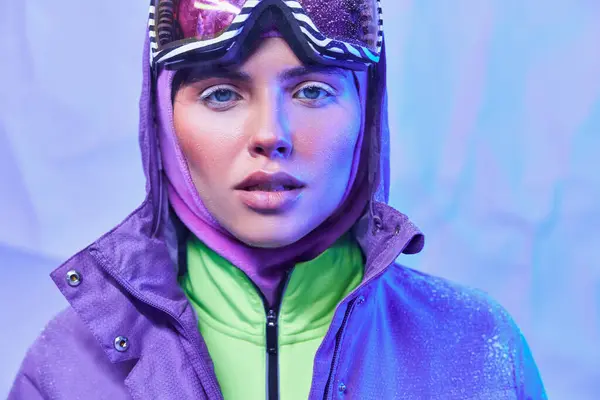 Aire helado, mujer joven en máscara de esquí, googles y chaqueta de invierno mirando a la cámara en el fondo azul - foto de stock