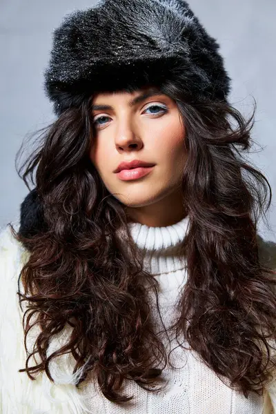 Брюнетка женщина в искусственной меховой шапке и белый свитер глядя на камеру на сером фоне, зимняя красота — стоковое фото