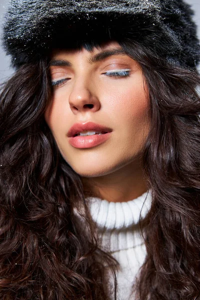 Кудрявая женщина в искусственной меховой шляпе и свитере позирует с закрытыми глазами на сером фоне, зимняя красота — стоковое фото