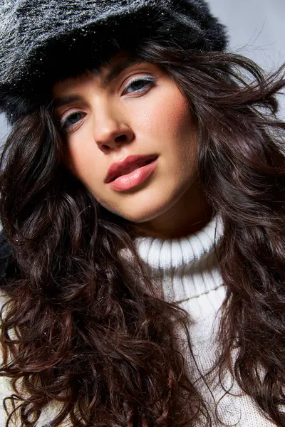 Красивая женщина в искусственной меховой шляпе и свитере, смотрящая в камеру на сером фоне, зимняя красота — стоковое фото