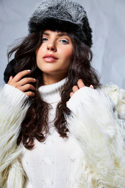 Красивая женщина в искусственной меховой куртке, шляпе и свитере, смотрящая в камеру на сером фоне, зимняя красота — стоковое фото