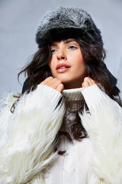 Очаровательная женщина в белой искусственной меховой куртке, шляпе и свитере позирует на сером фоне, зимняя красота — стоковое фото