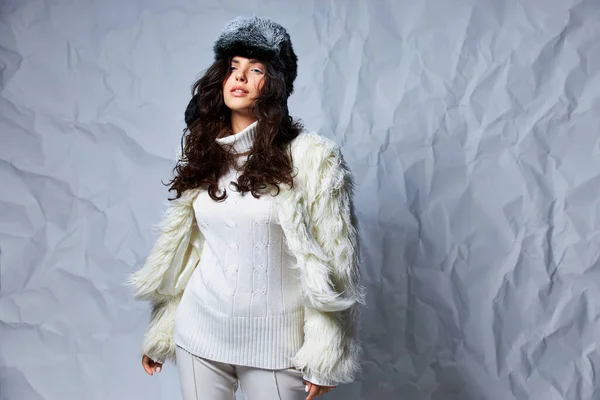 Великолепная кудрявая женщина в белом искусственном меховом пиджаке, зимней шляпе и свитере позирует на сером фоне — стоковое фото