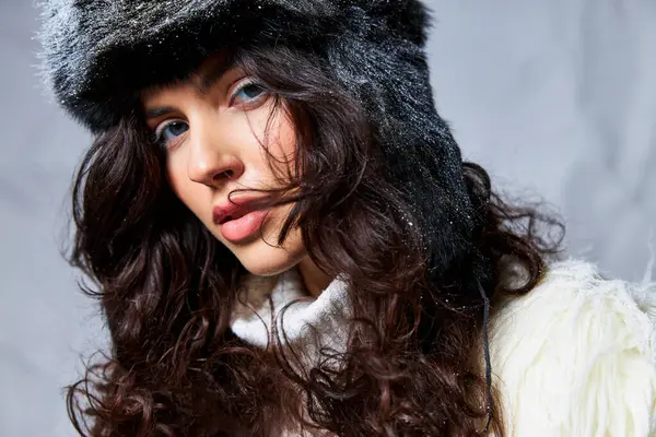 Snow on furry hat, curly brunette woman in white sweater looking at camera on grey backdrop — Stock Photo