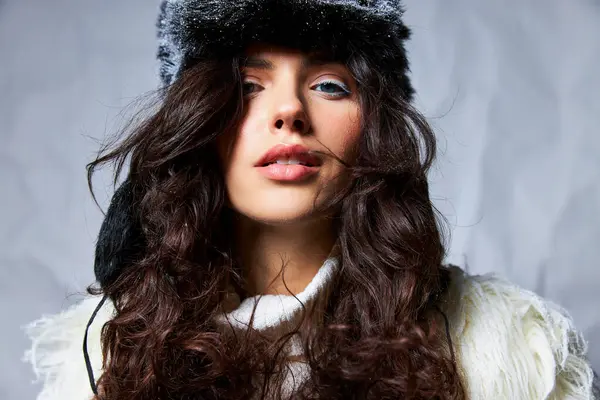 Snow queen, curly brunette woman in furry snowy hat and sweater looking at camera on grey backdrop — Stock Photo