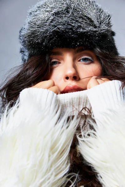 Atemberaubende Frau in weißer Kunstpelzjacke, Hut und Pullover posiert vor grauem Hintergrund, Wintermode — Stockfoto