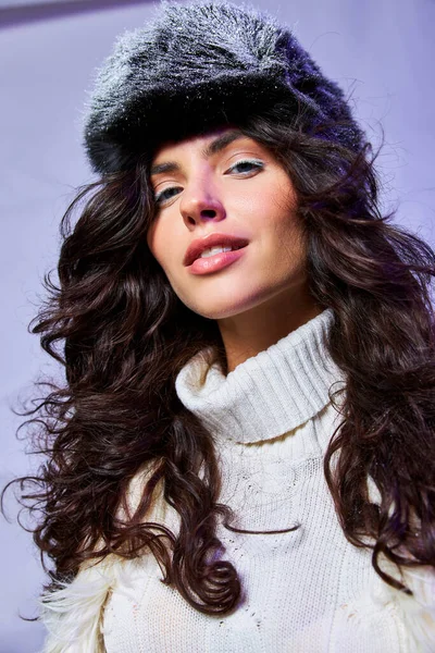 Mulher radiante em casaco de pele de falso branco, chapéu de inverno e suéter posando em pano de fundo cinza, olhos azuis — Fotografia de Stock