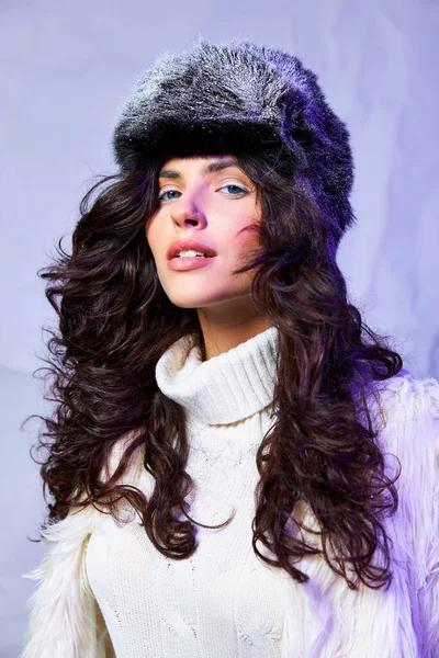 Morena mujer de ojos azules en chaqueta de piel sintética blanca, sombrero de invierno y suéter posando sobre fondo gris - foto de stock
