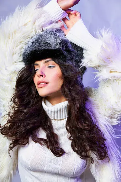 Curly brunette woman in faux fur jacket, winter hat and sweater posing with raised hand on blue — Stock Photo