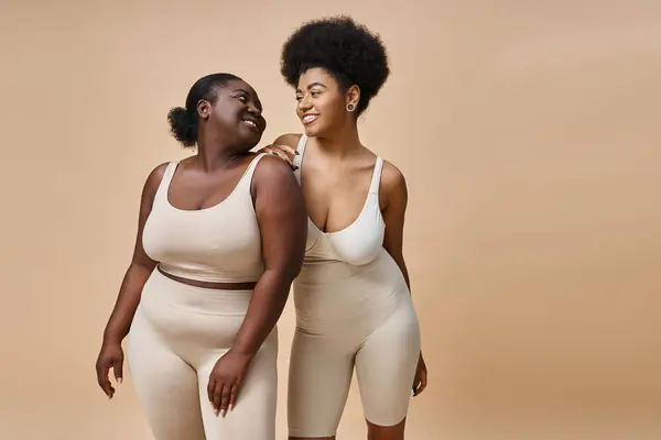 Счастливый плюс размер африканских американских женщин в нижнем белье улыбаясь друг другу на бежевый, тело позитивности — стоковое фото