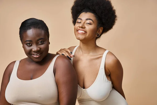 Счастливый плюс размер африканских американских женщин в нижнем белье улыбаясь на камеру на бежевый, тело позитивности — стоковое фото