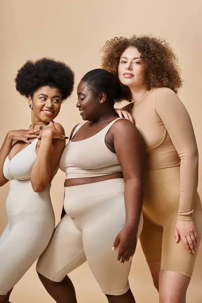 Fröhliche multiethnische Plus-Size-Frauen in Unterwäsche posieren vor beigem Hintergrund, Selbstbewusstsein und Charme — Stockfoto