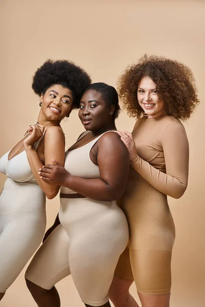 Happy multiracial plus size girlfriends in lingerie smiling on beige backdrop, body positive beauty — Stock Photo