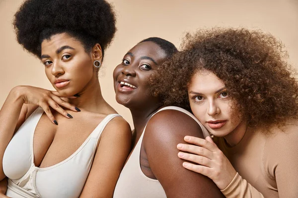 Cheerful plus size woman in lingerie looking at camera near multiethnic girlfriends on beige — Stock Photo