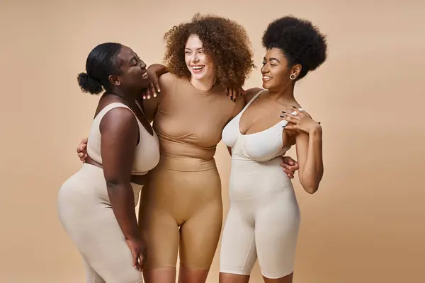 Feliz cuerpo multiétnico mujeres positivas en ropa interior abrazándose en beige, belleza curvilínea natural - foto de stock