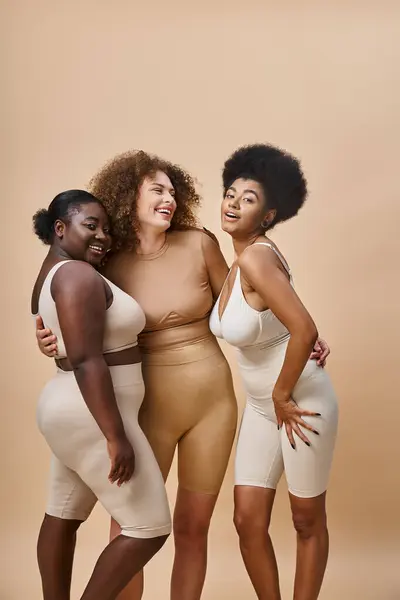 Feliz corpo multiétnico mulheres positivas em roupa interior abraçando em bege, beleza curvilínea natural — Fotografia de Stock