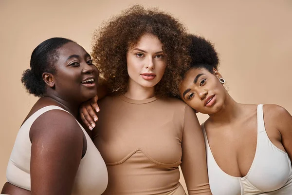 Expresivo cuerpo multiétnico mujeres positivas en lencería mirando a la cámara en beige, belleza natural - foto de stock