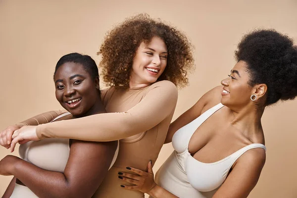 Alegre multicultural más tamaño de las mujeres en lencería abrazando en beige, positividad corporal y belleza - foto de stock
