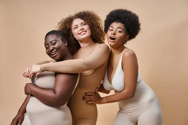 Cheerful multicultural plus size women in lingerie embracing on beige, body positivity and beauty — Stock Photo