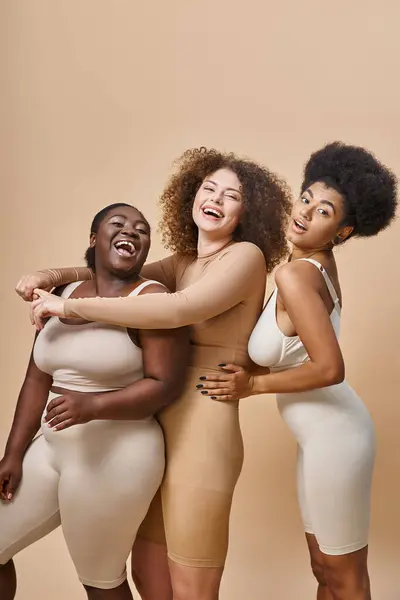 Emocionado multirracial más tamaño de las mujeres en lencería abrazando y riendo en beige, positividad corporal - foto de stock