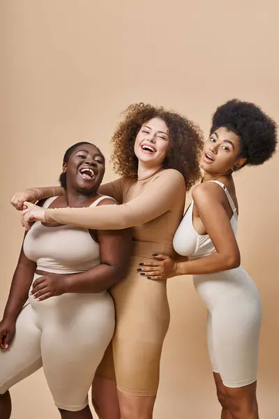 Excited multiracial plus size women in lingerie embracing and laughing on beige, body positivity — Stock Photo
