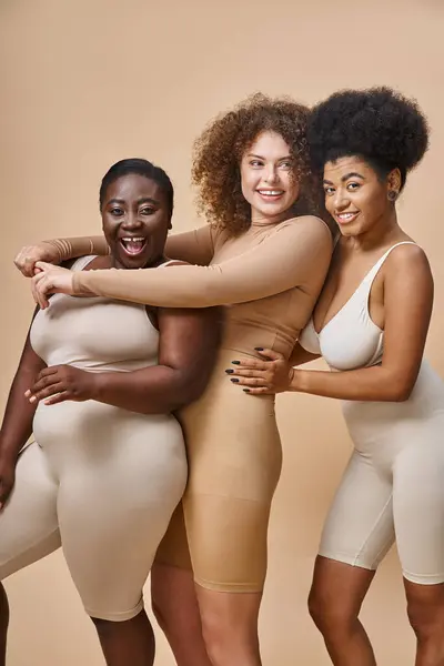 Emocionado multirracial más tamaño de las mujeres en lencería abrazando y riendo en beige, positividad corporal - foto de stock