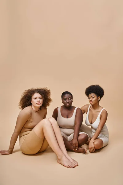 Curvy and body positive multiethnic women in lingerie sitting and looking at camera on beige — Stock Photo