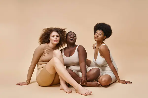 Curvas y el cuerpo positivo mujeres multiétnicas en lencería sentado y mirando a la cámara en color beige - foto de stock