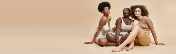 Curvy plus size mulheres multiétnicas em lingerie sentado e olhando para a câmera em bege, banner — Fotografia de Stock