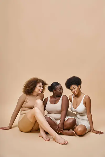 Full length of multicultural plus size girlfriends in underwear sitting on beige, self-expression — Stock Photo