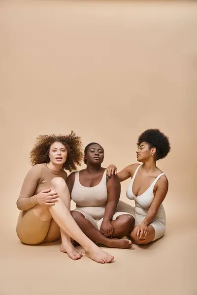 Full length of multicultural plus size girlfriends in underwear sitting on beige, self-expression — Stock Photo