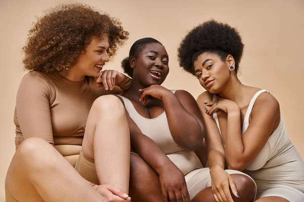 Despreocupado africano americano más tamaño mujer en lencería riendo cerca multiétnica novias en beige - foto de stock