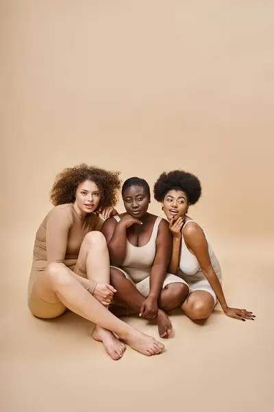 Радісне багатоетнічне тіло позитивні жінки в нижній білизні сидять і посміхаються на бежевій, самооцінці — стокове фото
