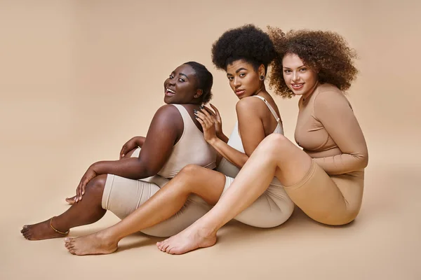 Fröhliche multiethnische körperpositive Frauen in Dessous sitzen und lächeln auf beige, Selbstwertgefühl — Stockfoto