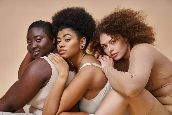 Ausdrucksstarke körperpositive Frauen in Unterwäsche posieren und in die Kamera schauen auf beige, Selbstbewusstsein — Stockfoto